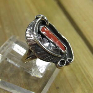 May include: A silver ring with a red coral stone set in a detailed design. The ring is on a clear display stand.