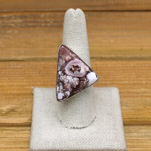 May include: A triangular ring featuring a large, patterned stone. The stone displays a mix of burgundy, white, and light pink hues, set in a silver-toned band. The ring is displayed on a neutral-colored ring holder, showcasing its unique design.