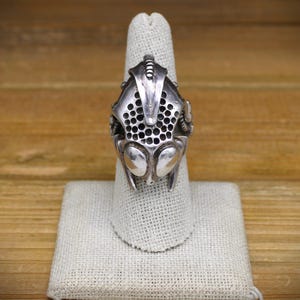 May include: A silver ring with a detailed design of a bug's head. The ring is on a white fabric ring holder.