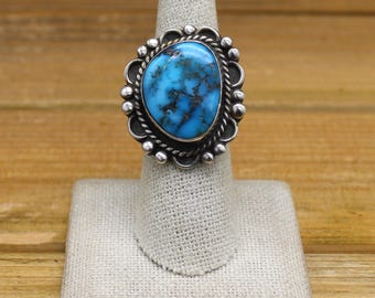 May include: A silver ring with a blue turquoise stone set in a decorative silver bezel. The ring is on a white ring display stand.