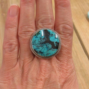 May include: A turquoise and silver ring with a round turquoise stone set in a silver bezel. The stone has a marbled pattern of blue and black.
