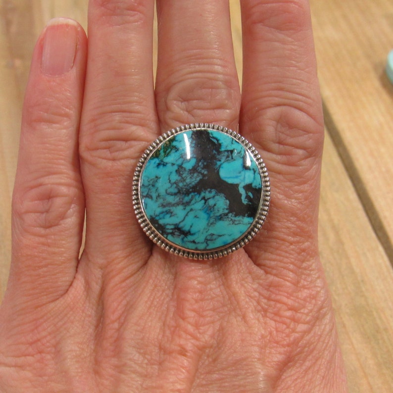 May include: A turquoise and silver ring with a round turquoise stone set in a silver bezel. The stone has a marbled pattern of blue and black.