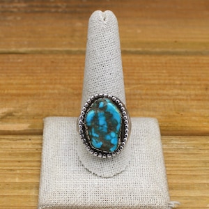 May include: A silver ring featuring a large, oval turquoise stone with dark matrix patterns. The stone is set in a bezel with a beaded border. The ring is displayed on a white fabric ring holder against a wooden background.