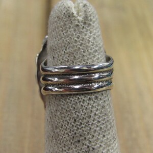 May include: Silver ring with three bands, each with a textured surface. The ring is on a fabric display stand.