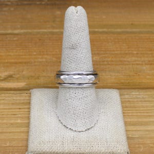 May include: A silver-colored ring with a hammered texture and a spinning band. The ring is displayed on a white fabric finger stand against a wooden background. The ring's design features a textured center band and smooth outer bands.