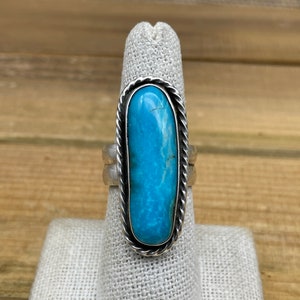 May include: A silver ring with a blue turquoise stone set in a rope design. The ring is on a white fabric ring holder.