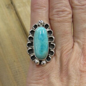 May include: A silver ring with a turquoise stone set in a decorative oval bezel. The stone is a light blue color with darker blue veins.