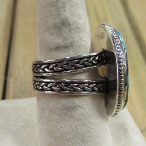 May include: A silver ring with a braided band and a turquoise stone set in a silver bezel. The stone is oval shaped and has a dark blue and green color.