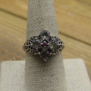 May include: A silver ring with a cluster of five clear stones and a single red stone in the center. The ring has a filigree design.