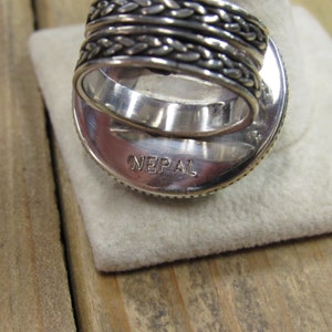 May include: Silver ring with a braided band and a round, textured top. The ring is stamped with the word "NEPAL" on the bottom.