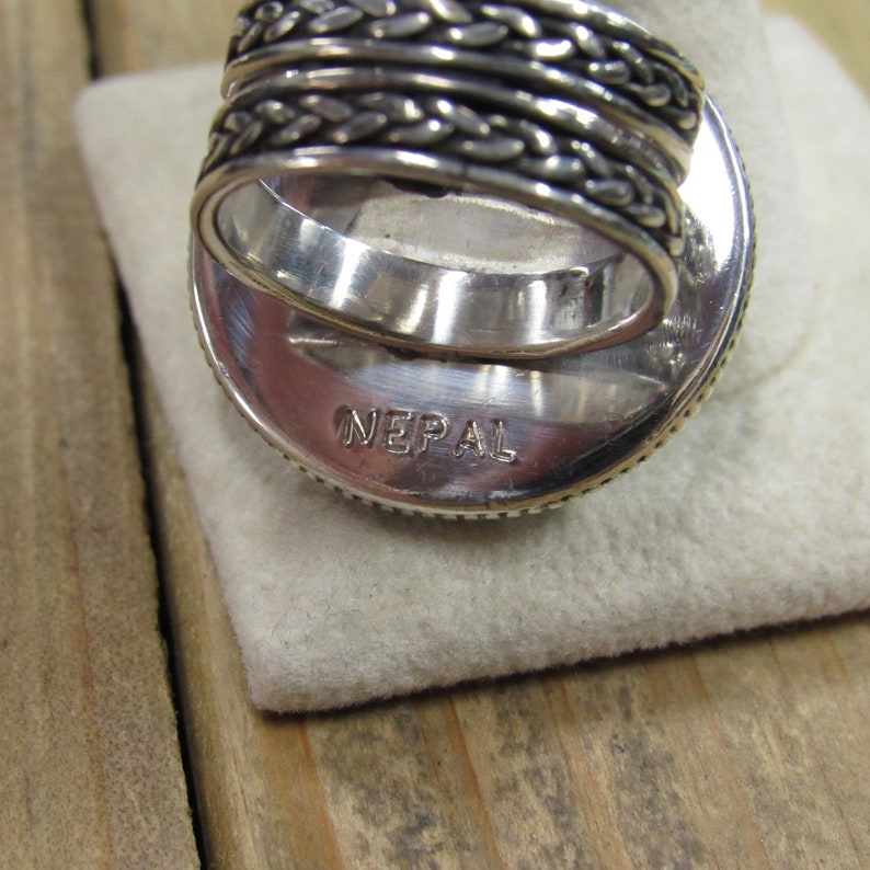 May include: Silver ring with a braided band and a round, textured top. The ring is stamped with the word "NEPAL" on the bottom.