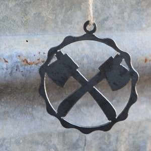 May include: A metal ornament shaped like two crossed axes, hanging from a twine string. The ornament is a dark gray color and has a scalloped edge.