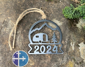 Könnte beinhalten: Ein Weihnachtsbaumschmuck aus Metall in Form eines Wohnwagens vor einer Bergszene. Der Schmuck hat ein Banner mit dem Jahr 2024 darauf.