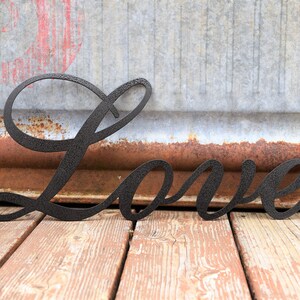 May include: Black metal word "Love" in a cursive font. The word is sitting on a wooden surface.