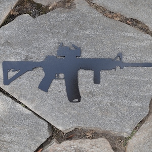 May include: Black metal silhouette of an assault rifle with a red dot sight on a gray stone background.