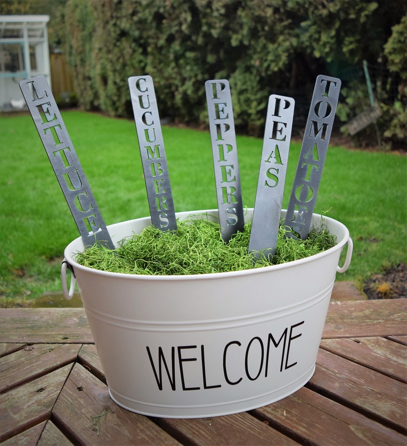 May include: Five metal garden markers with the words "Lettuce", "Cucumbers", "Peppers", "Peas", and "Tomato" are displayed in a white planter with the word "Welcome" on the side. The planter is filled with green moss.