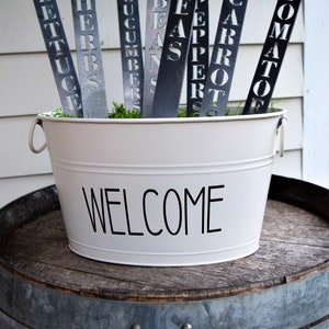 May include: A white metal bucket with the word "WELCOME" printed on the front. The bucket is filled with green moss and metal garden markers with the words "LETTUCE", "HERBS", "CUCUMBERS", "BEANS", "PEPPER", "CARROT", and "TOMATO" printed on them.