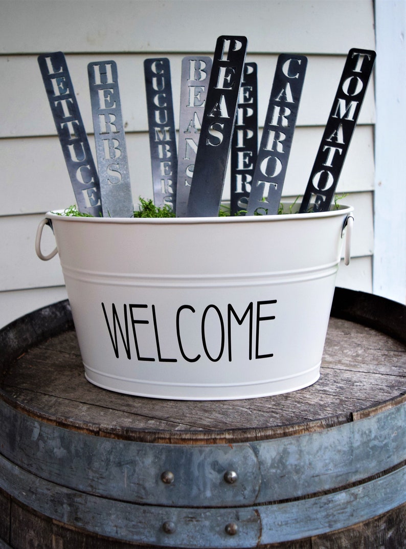 May include: A white metal bucket with the word "WELCOME" printed on the front. The bucket is filled with green moss and metal garden markers with the words "LETTUCE", "HERBS", "CUCUMBERS", "BEANS", "PEPPER", "CARROT", and "TOMATO" printed on them.