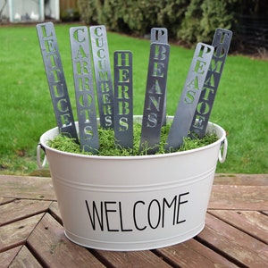 May include: A white metal bucket with the word "WELCOME" printed on the front. The bucket is filled with green moss and contains several metal plant markers with the words "LETTUCE", "CARROTS", "CUCUMBERS", "HERBS", "BEANS", "PEAS", and "TOMATOES" cut out.