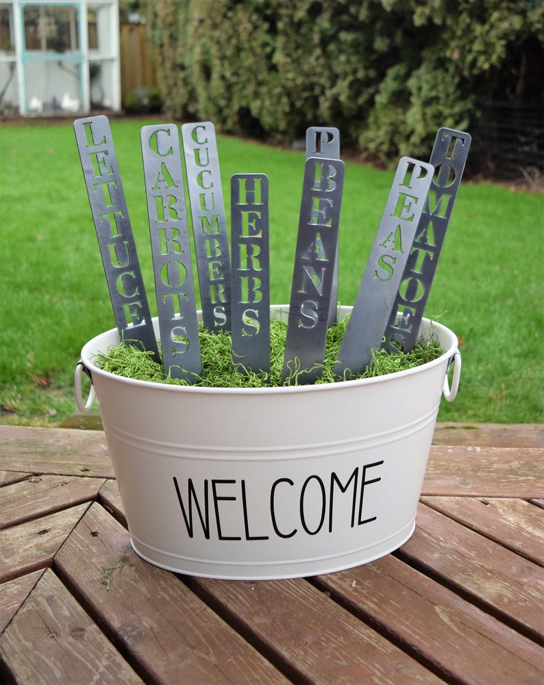 May include: A white metal bucket with the word "WELCOME" printed on the front. The bucket is filled with green moss and contains several metal plant markers with the words "LETTUCE", "CARROTS", "CUCUMBERS", "HERBS", "BEANS", "PEAS", and "TOMATOES" cut out.