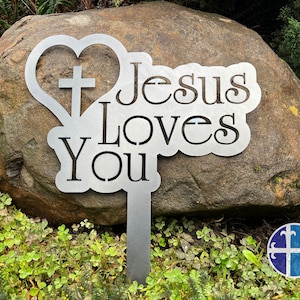 Puede incluir: Estaca de jard&iacute;n de metal con forma de coraz&oacute;n y cruz, y las palabras "Jes&uacute;s te ama".