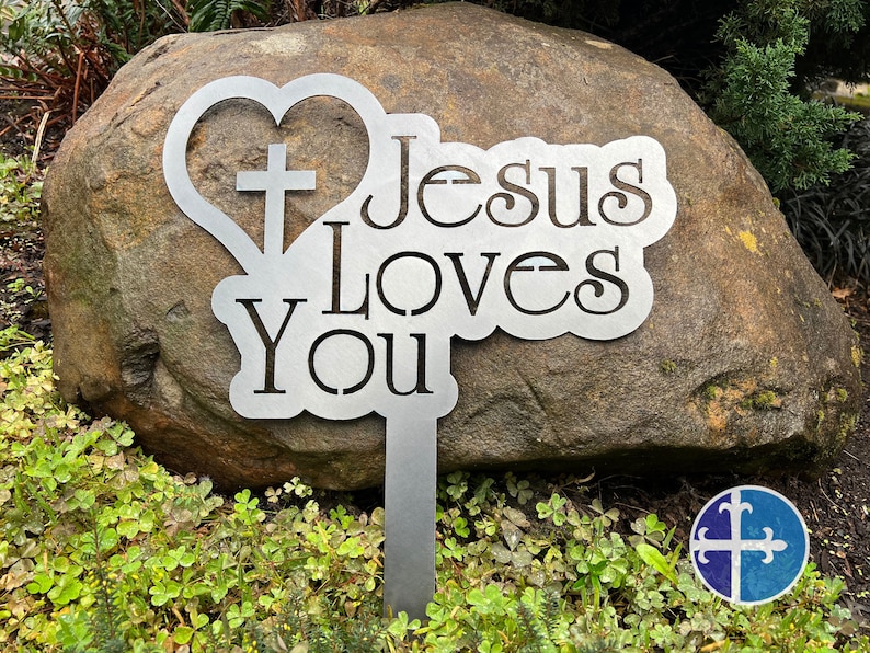 Puede incluir: Estaca de jard&iacute;n de metal con forma de coraz&oacute;n y cruz, y las palabras "Jes&uacute;s te ama".
