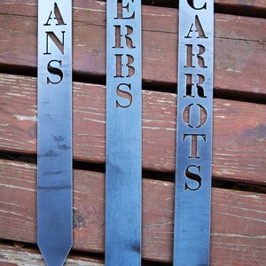 May include: Three metal garden markers with pointed ends. The markers are labelled "BEANS", "HERBS", and "CARROTS" in a simple, blocky font.