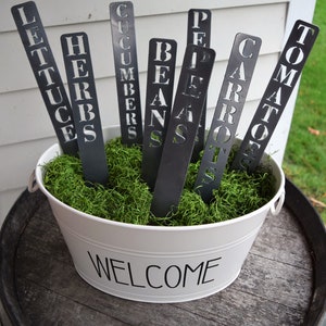 May include: A white metal bucket with the word "WELCOME" printed on the front. The bucket is filled with green moss and contains nine metal garden markers with the names of different vegetables: LETTUCE, HERBS, CUCUMBERS, BEANS, PEPPERS, CARROTS, TOMATOES.