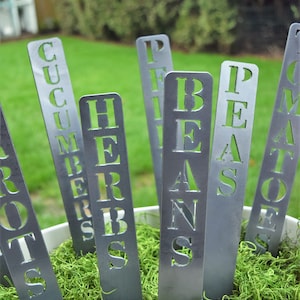 May include: Metal garden markers with the words "Tomatoes", "Peas", "Beans", "Herbs", "Cucumbers", and "Potatoes" written on them. The markers are placed in a pot with green moss.