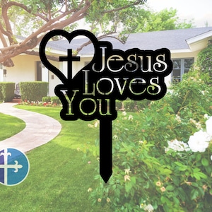 Puede incluir: Se&ntilde;al de jard&iacute;n de metal negro con forma de coraz&oacute;n que contiene una cruz y las palabras "Jes&uacute;s te ama". La se&ntilde;al est&aacute; delante de un c&eacute;sped verde y una casa.