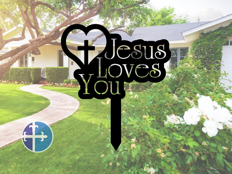Puede incluir: Se&ntilde;al de jard&iacute;n de metal negro con forma de coraz&oacute;n que contiene una cruz y las palabras "Jes&uacute;s te ama". La se&ntilde;al est&aacute; delante de un c&eacute;sped verde y una casa.