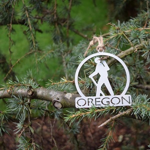 May include: A silver metal ornament featuring a hiker silhouette inside a circle with the word "OREGON" below. The ornament is hanging from a branch of a coniferous tree.
