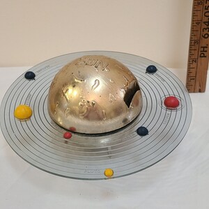 May include: A silver model of the solar system with a large, round, silver sun in the center and smaller, colored planets orbiting around it. The planets are made of plastic and are red, yellow, and blue.