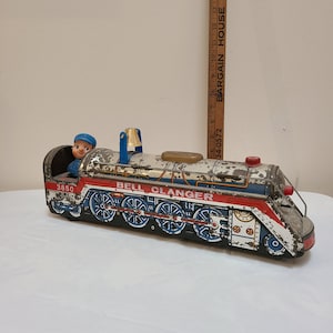Puede incluir: Tren de juguete de hojalata vintage con una locomotora azul y roja, una campana en la parte superior y una pequeña figura de un conductor en la cabina. El tren está etiquetado como "BELL CLANGER" y "3850".