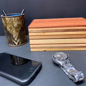 May include: A wooden box with a layered wood design. The box has a dark red top and a light wood base. The box is sitting on a black surface next to a cell phone and a silver watch.