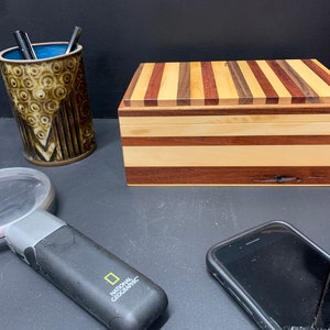 May include: A wooden box with a striped lid made of different types of wood. The box is closed and sitting on a dark surface. There is a magnifying glass and a pencil holder with pencils next to the box.