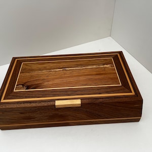 May include: A rectangular wooden box with a hinged lid. The box is made of dark brown wood with lighter wood accents and a rectangular inlay on the lid. A small, light-coloured wooden latch secures the box.