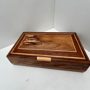 May include: A rectangular wooden box with a hinged lid. The box is made of dark brown wood with a lighter wood trim and a light-coloured wooden latch. The top of the box features a decorative wood grain pattern.