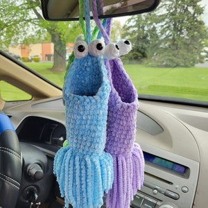 May include: Two crocheted hanging ornaments with googly eyes and long fringe. The ornaments are blue and purple and are hanging from a rear view mirror in a car.