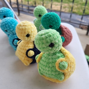 Könnte beinhalten: Eine Sammlung von gehäkelten Schildkrötenspielzeugen in verschiedenen Farben, darunter Grün, Gelb, Blau und Türkis. Die Schildkröten haben schwarze Augen und sind aus weichem, flauschigem Garn gefertigt.