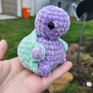 May include: A crocheted plush toy shaped like a turtle. The turtle is purple and green with a black eye.