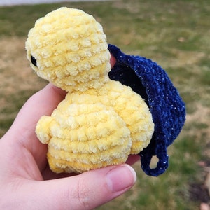 May include: A yellow crocheted plush toy with a blue crocheted cape. The toy is shaped like a duck and has a small, round head with black eyes.