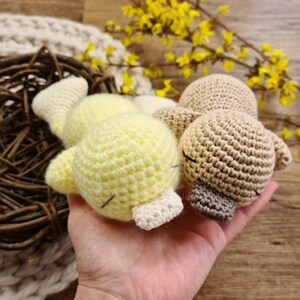 May include: Two crocheted stuffed birds, one yellow and one beige, are held in a hand. The birds are lying down with their heads together.
