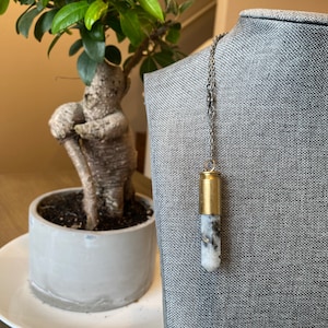 Puede incluir: Un collar de cadena plateada con un colgante de latón en forma de casquillo de bala relleno de una piedra blanca y negra.