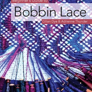以下が含まれることがあります： Gilian DyeとAdrienne Thunderによる「Beginner's Guide to Bobbin Lace」というタイトルの本。表紙には、ドット柄の作業台の上で、多数の木製ボビンと糸を使って作られている、青と紫の繊細なレース模様が描かれています。