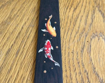 May include: A dark wooden bookmark featuring two koi fish illustrations. One koi is orange, and the other is red and white. The bookmark has a gold tassel attached at the top. Small gold dots are scattered around the fish.