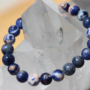 Peut inclure: Un bracelet en pierres précieuses bleues et blanches avec des perles rondes. Le bracelet est fait de pierres naturelles et est présenté sur un fond de cristal blanc.