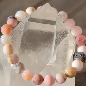 Peut inclure: Un bracelet en perles d'opale rose avec un charme argenté. Le bracelet est sur un fond de cristal blanc.