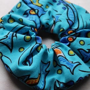 May include: A turquoise blue scrunchie with a colorful fish pattern. The fish are blue, yellow, orange, and green. The scrunchie is made of fabric and has a gathered look.