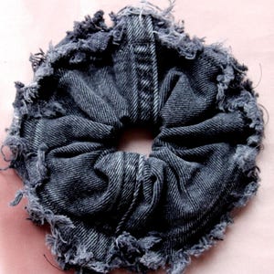 Puede incluir: Una goma para el pelo de mezclilla gris oscuro con bordes deshilachados. La tela se junta alrededor de una abertura central, creando un accesorio circular texturizado. Esta cinta para el pelo hecha a mano añade un toque de estilo casual.